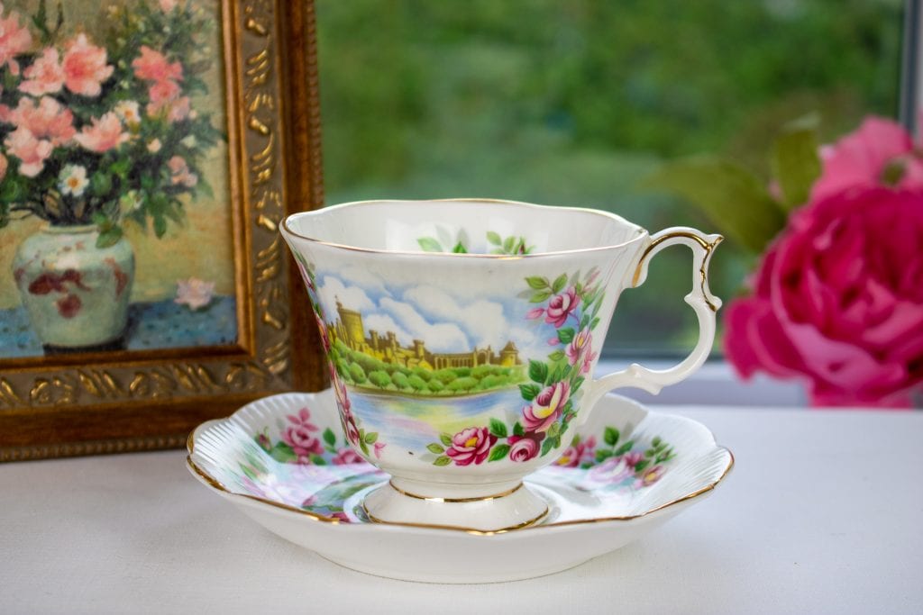 A Royal Albert teacup and saucer sits on a table with a white table cloth in front of a window. In the background, there are pink peonies to the right and a painting in a small vintage frame to the left.