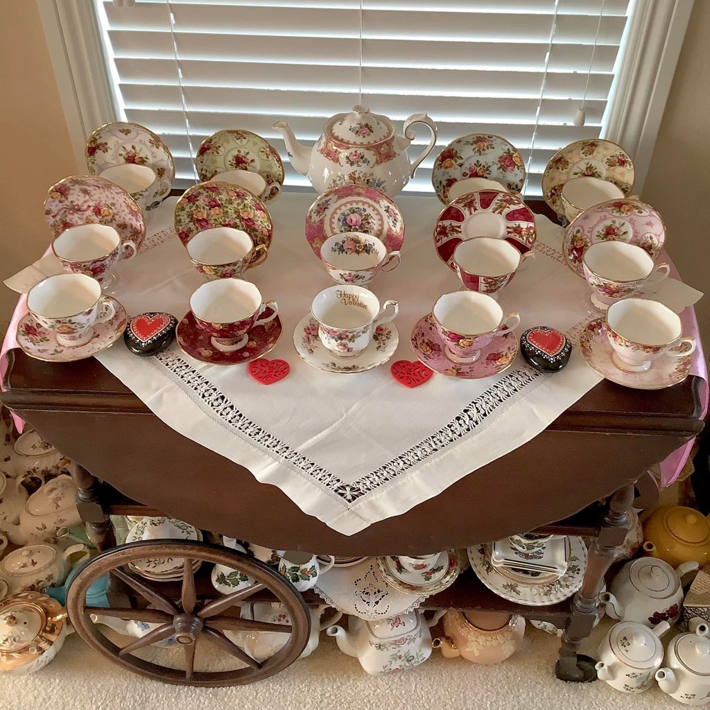 A small white table cloth is draped over a brown wood tea wagon with different pink and red Royal Albert teacups on it in 3 rows.